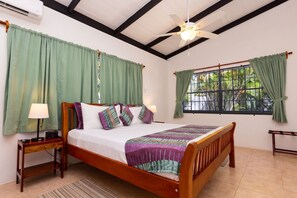 A bedroom with a wooden bed, green curtains, a ceiling fan, and a window with a view of greenery. - Lance Aux Epines Cottages (St. George's)