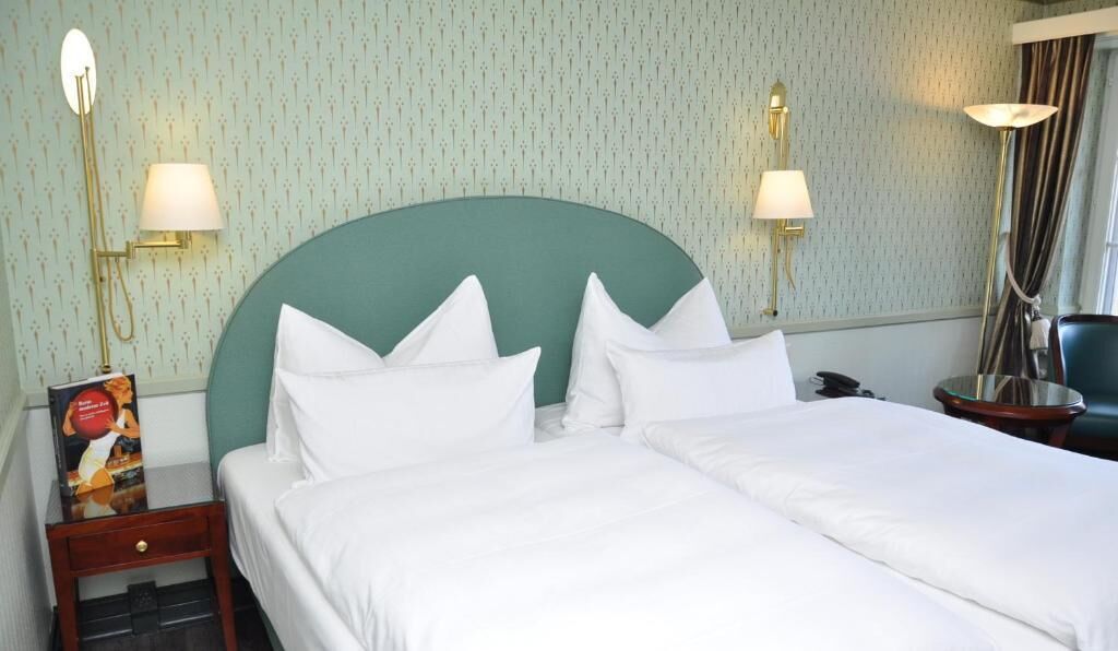 Habitación de hotel con una cama grande, dos lámparas de noche, una mesita de noche con un libro y un cabecero verde.