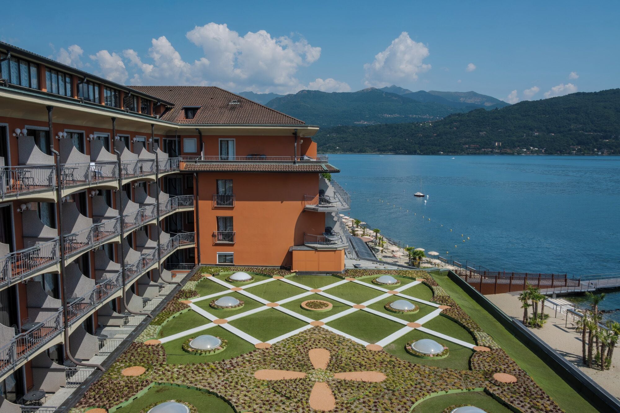 Un hotel con terraza que presenta un diseño de jardín geométrico, una piscina y vistas a un lago con montañas al fondo.