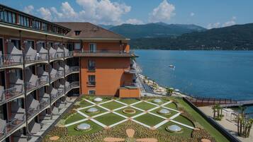 Un hotel con terraza que presenta un diseño de jardín geométrico, una piscina y vistas a un lago con montañas al fondo.