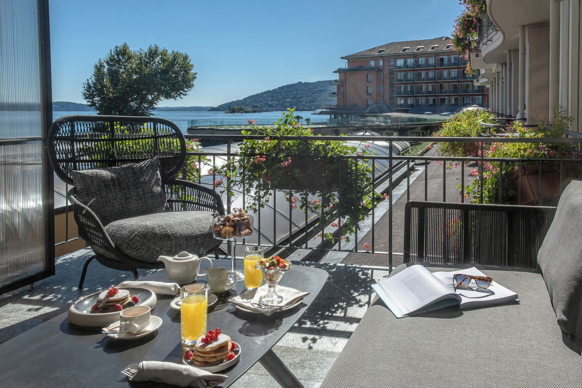 A balcony with wicker furniture, a table set with breakfast items, and a view of a lake and buildings.