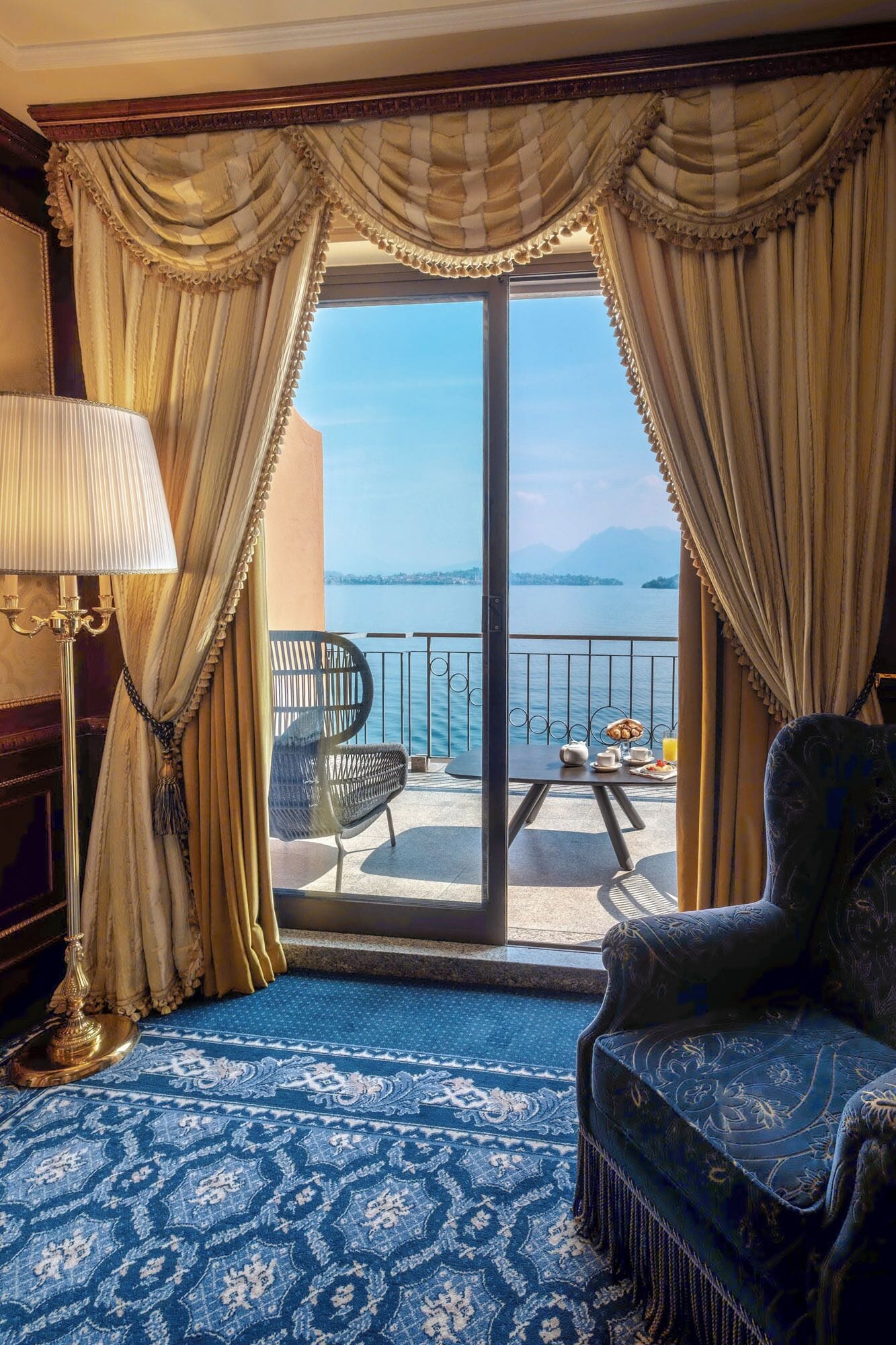 Una stanza con vista sul mare, caratterizzata da un tappeto azzurro a motivi decorativi, un divano, una poltrona e un tavolo imbandito per la colazione.