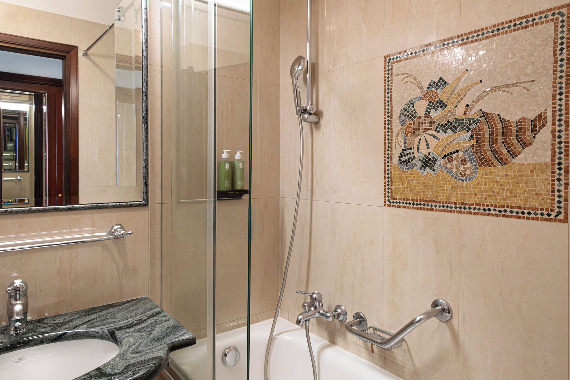 A bathroom with a glass shower enclosure, a bathtub with a grab bar, a sink with a marble countertop, and a mosaic tile wall.