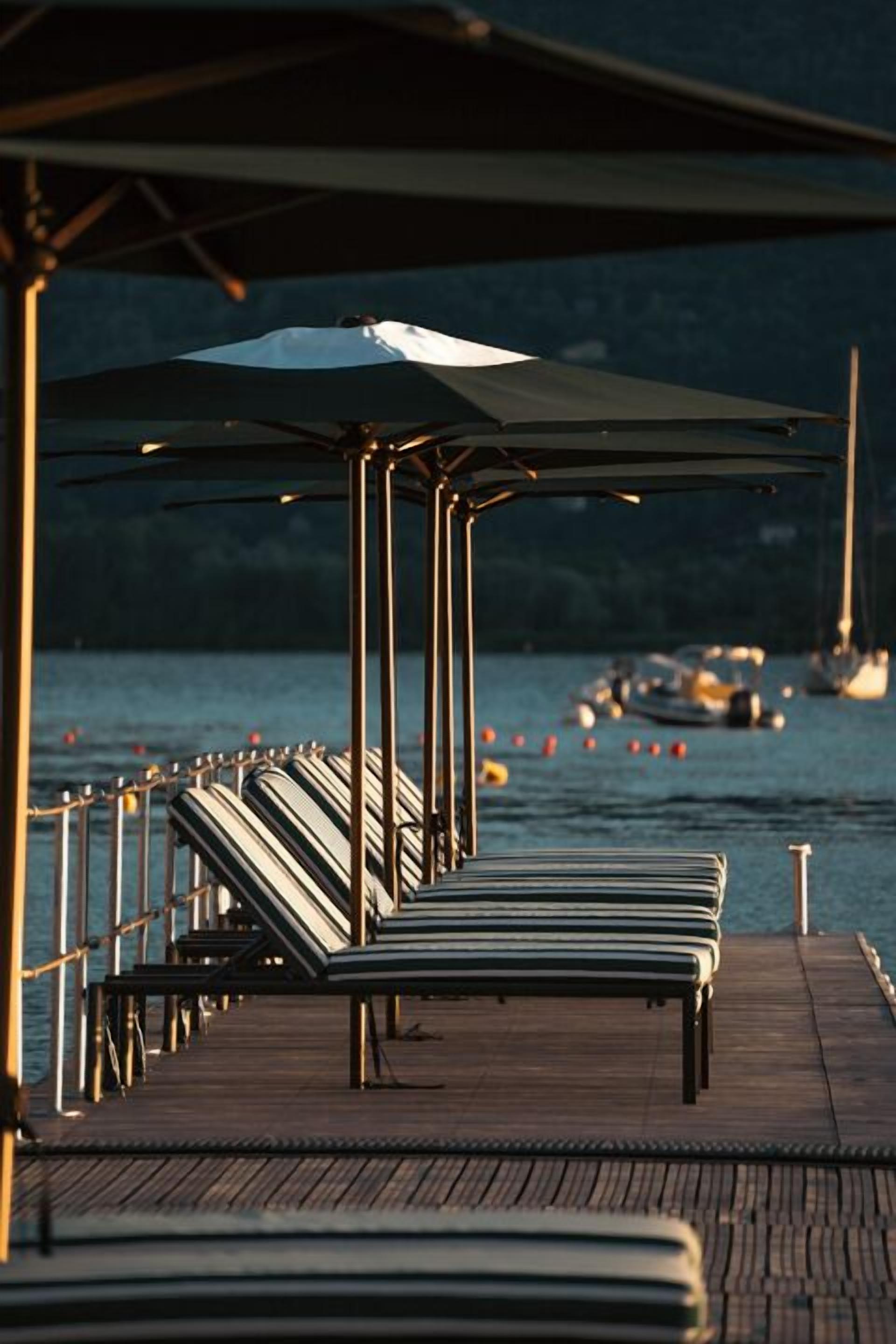 A wooden pier with striped lounge chairs and umbrellas, overlooking calm water with boats in the distance.