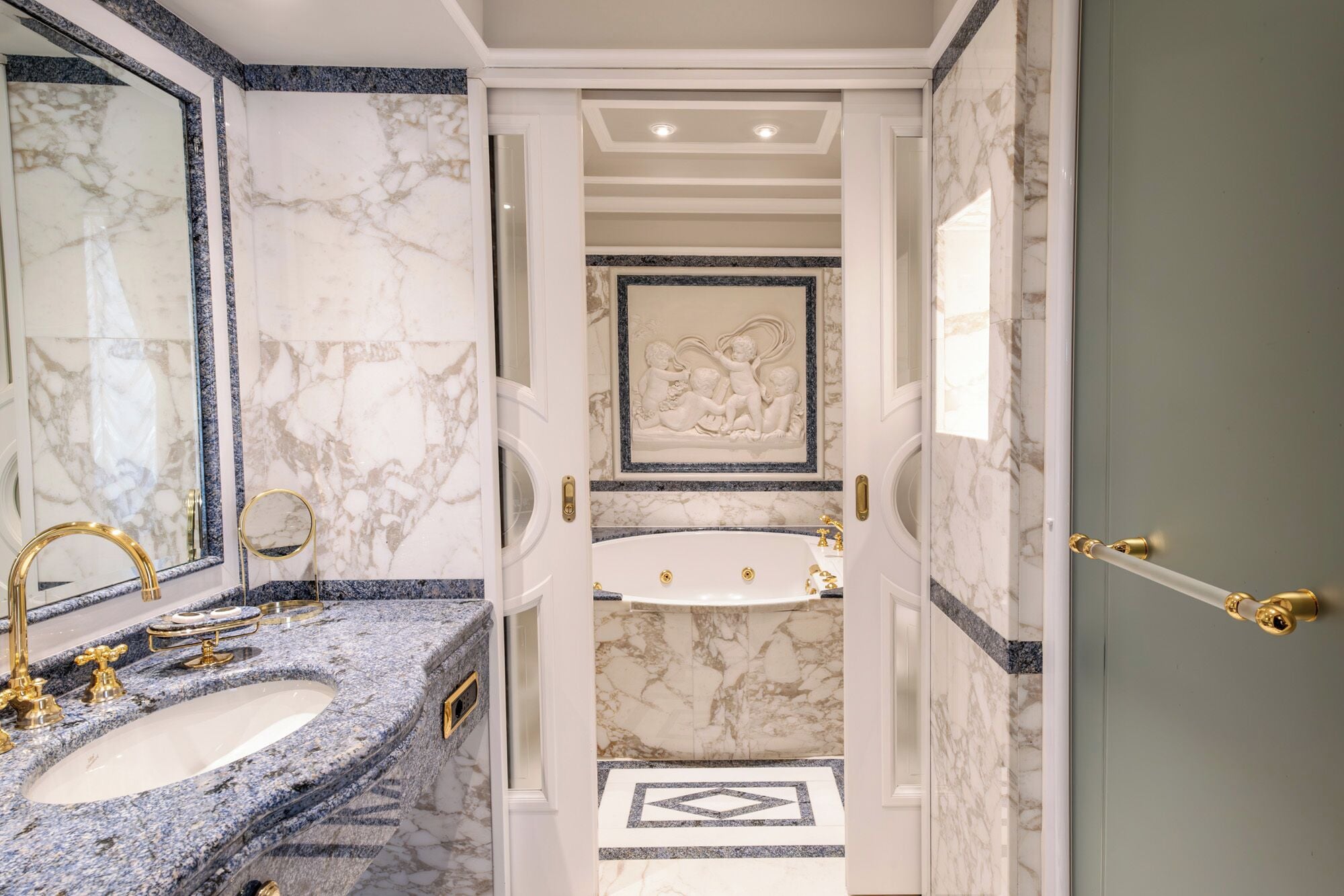 Ein luxuriöses Badezimmer mit Marmorwänden, einem Doppelwaschbecken mit goldfarbenen Armaturen und einer dekorativen Reliefdarstellung über dem Waschbecken.