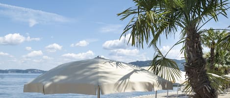 A beach with sun loungers and umbrellas, palm trees, and a clear view of the sea.