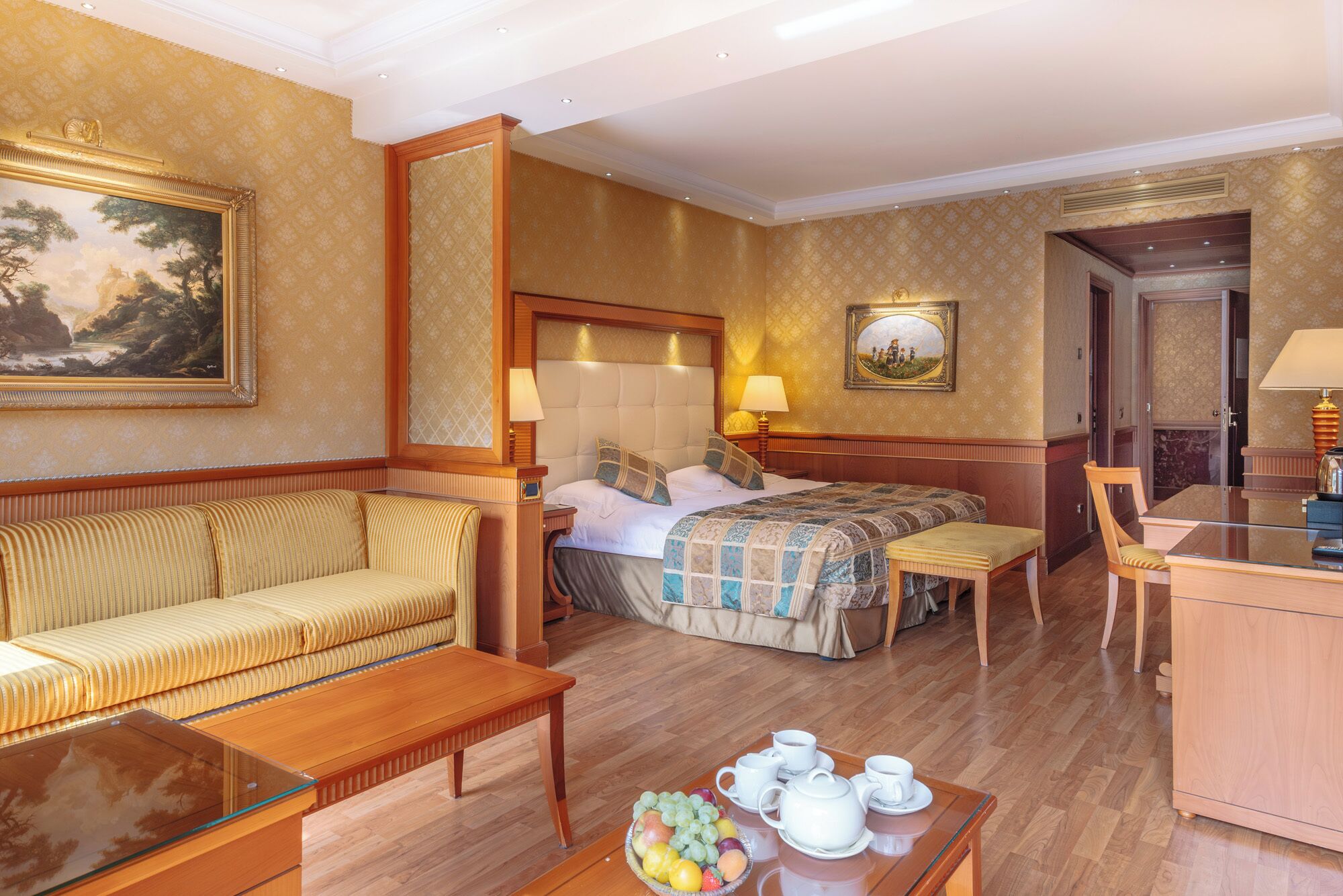 A hotel room with a bed, a sofa, a dining table with chairs, a small table with a fruit bowl and tea set, and a painting on the wall.