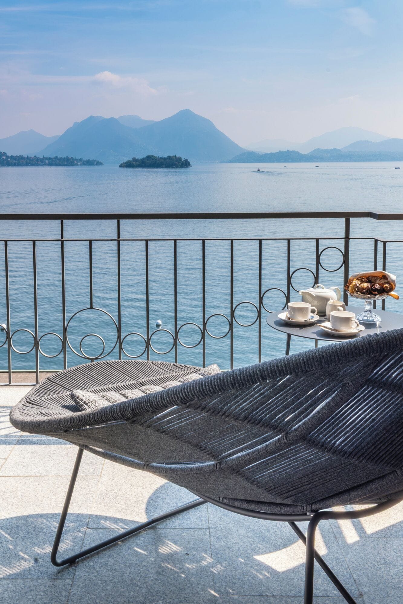 A balcony with a wicker chair, a table with a teapot, cups, and a dessert bowl.