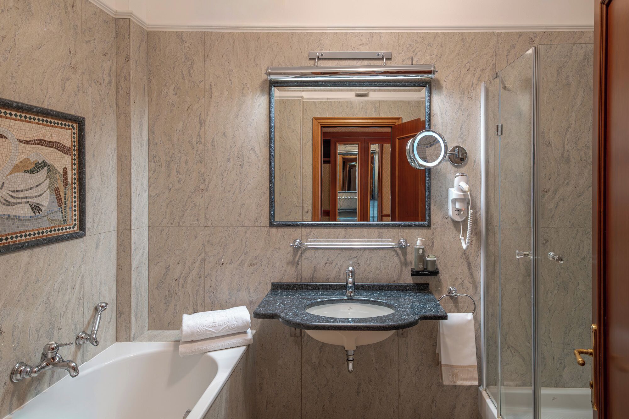 A bathroom with a marble wall, a sink, a bathtub, a glass shower enclosure, a mirror, and a towel rack.