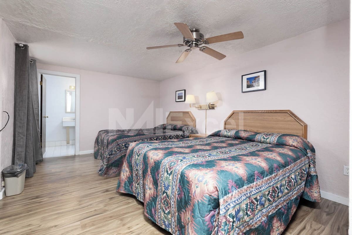 A hotel room with two beds, a ceiling fan, a bathroom visible through an open door, and a nightstand with a lamp.