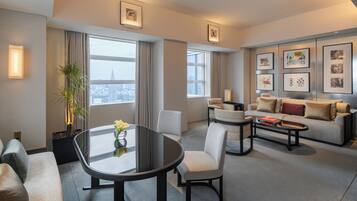 A modern hotel room with a dining area, a sofa set, a coffee table, and a large window offering a city view.