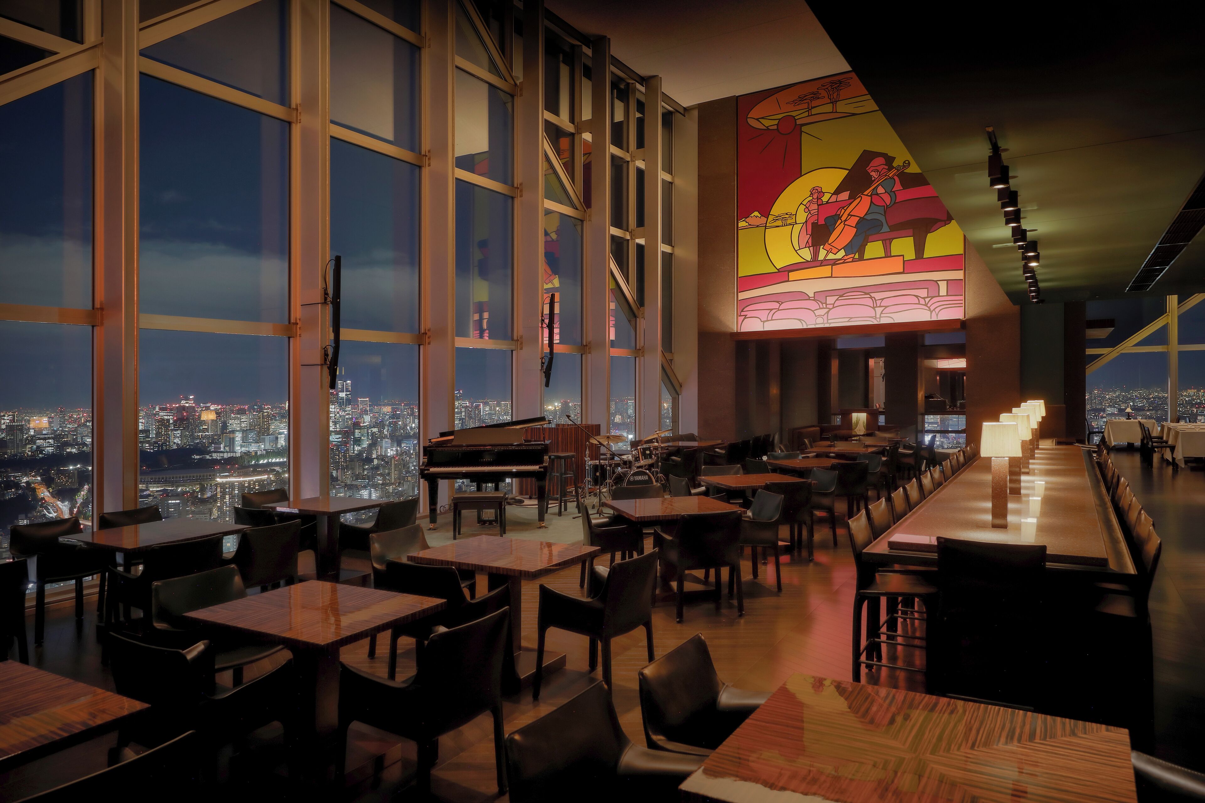 A restaurant with large windows offering a city view, a piano, and a mural on the wall.