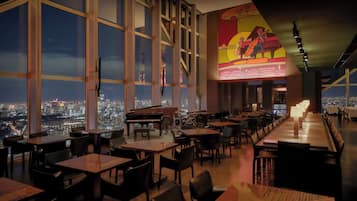 A restaurant with large windows offering a city view, a piano, and a mural on the wall.