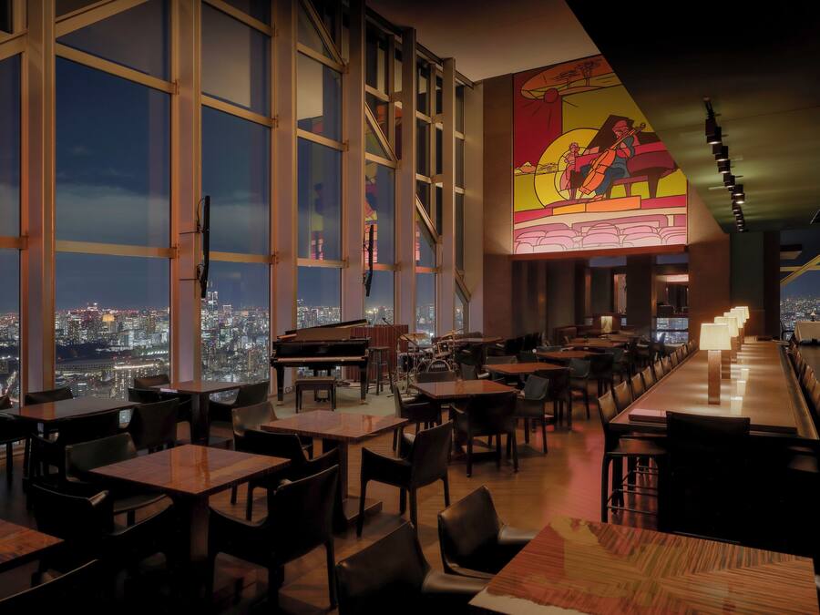 A restaurant with large windows offering a city view, a piano, and a mural on the wall.
