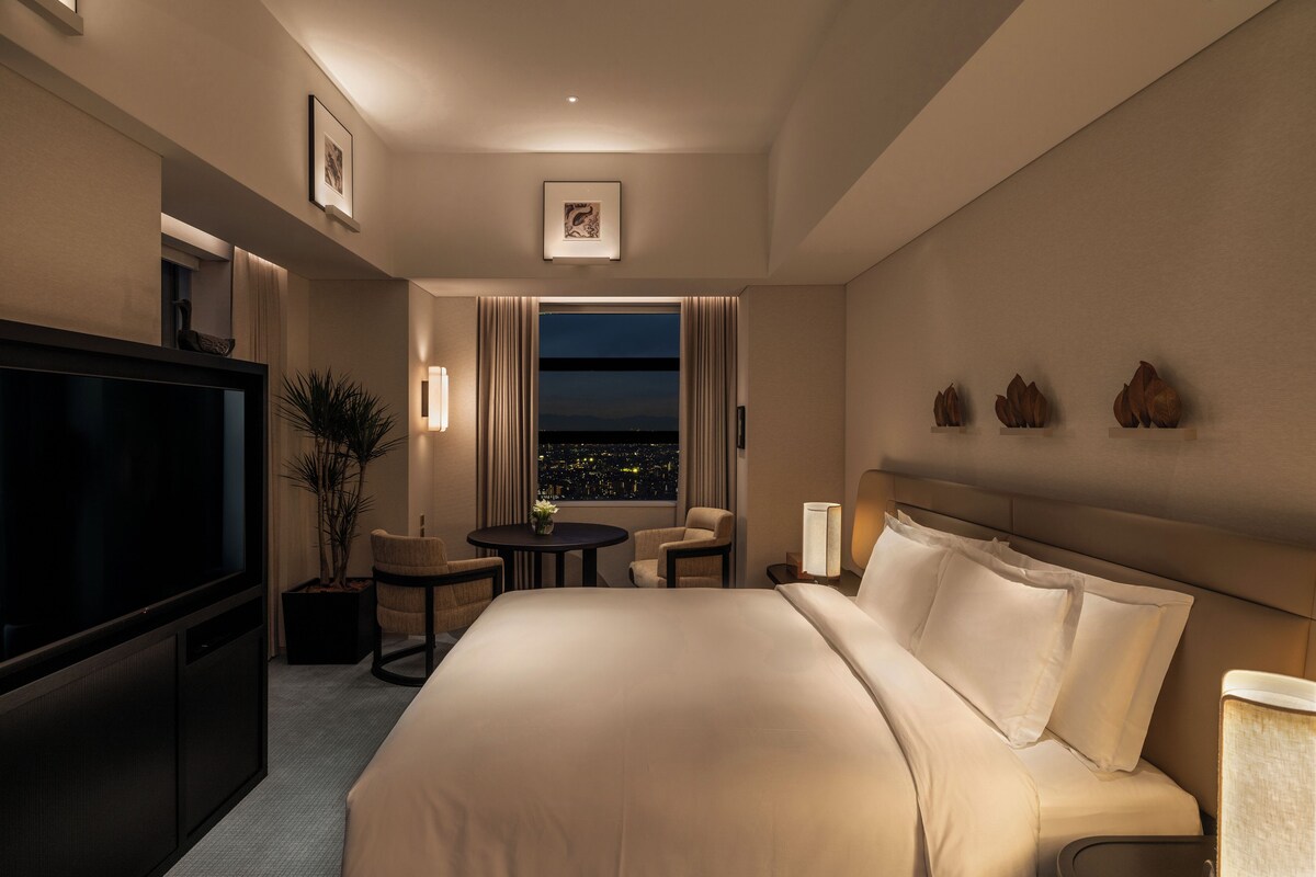 Habitación de hotel con una cama grande, televisión, una pequeña mesa con sillas, una planta en maceta y vista nocturna de la ciudad.