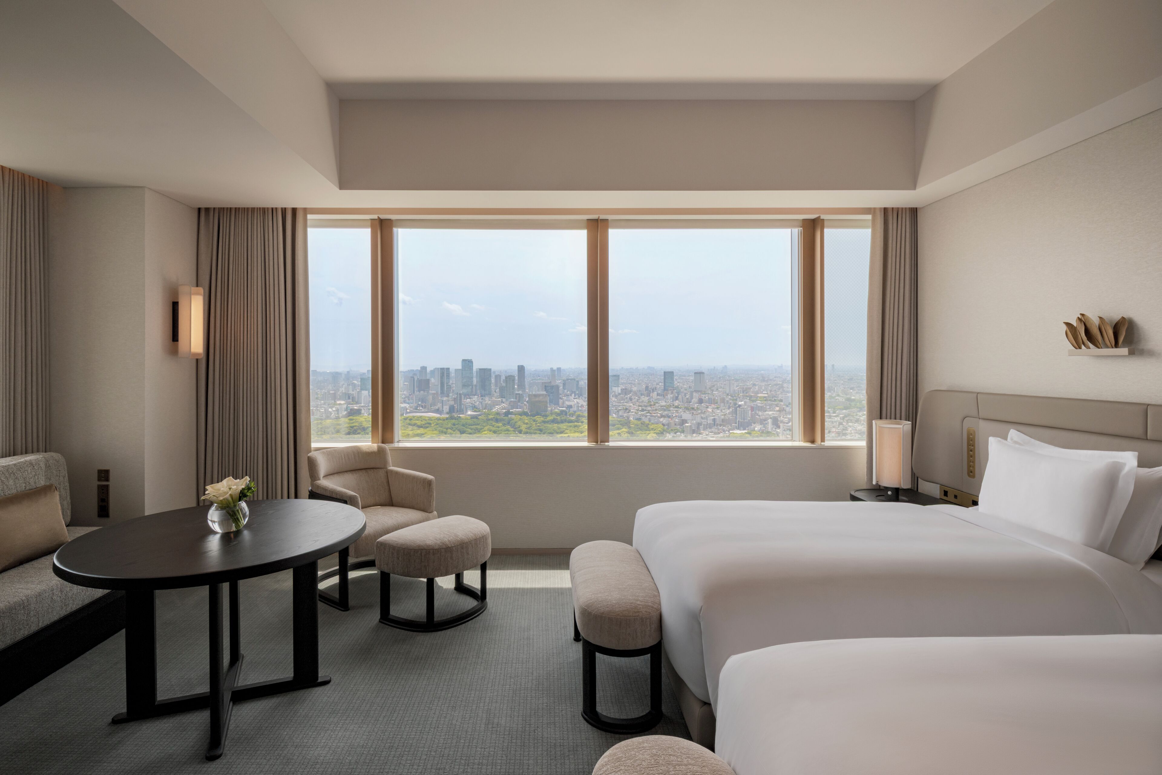 Habitación de hotel con una cama grande, un sofá pequeño, una mesa redonda y vistas a un paisaje urbano a través de amplios ventanales.