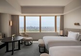 A hotel room with a large bed, a smaller sofa, a round table, and a view of a cityscape through large windows.