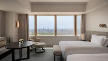 Habitación de hotel con una cama grande, un sofá pequeño, una mesa redonda y vistas a un paisaje urbano a través de amplios ventanales.
