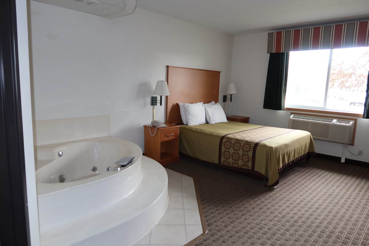 Una habitación de hotel con una bañera grande, una cama con cabecero, dos lámparas de noche y un ventanal con cortinas.