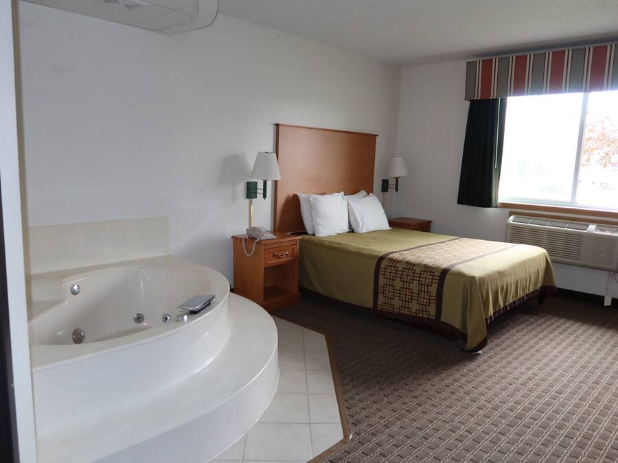 A hotel room with a large bathtub, a bed with a headboard, two bedside lamps, and a window with curtains.