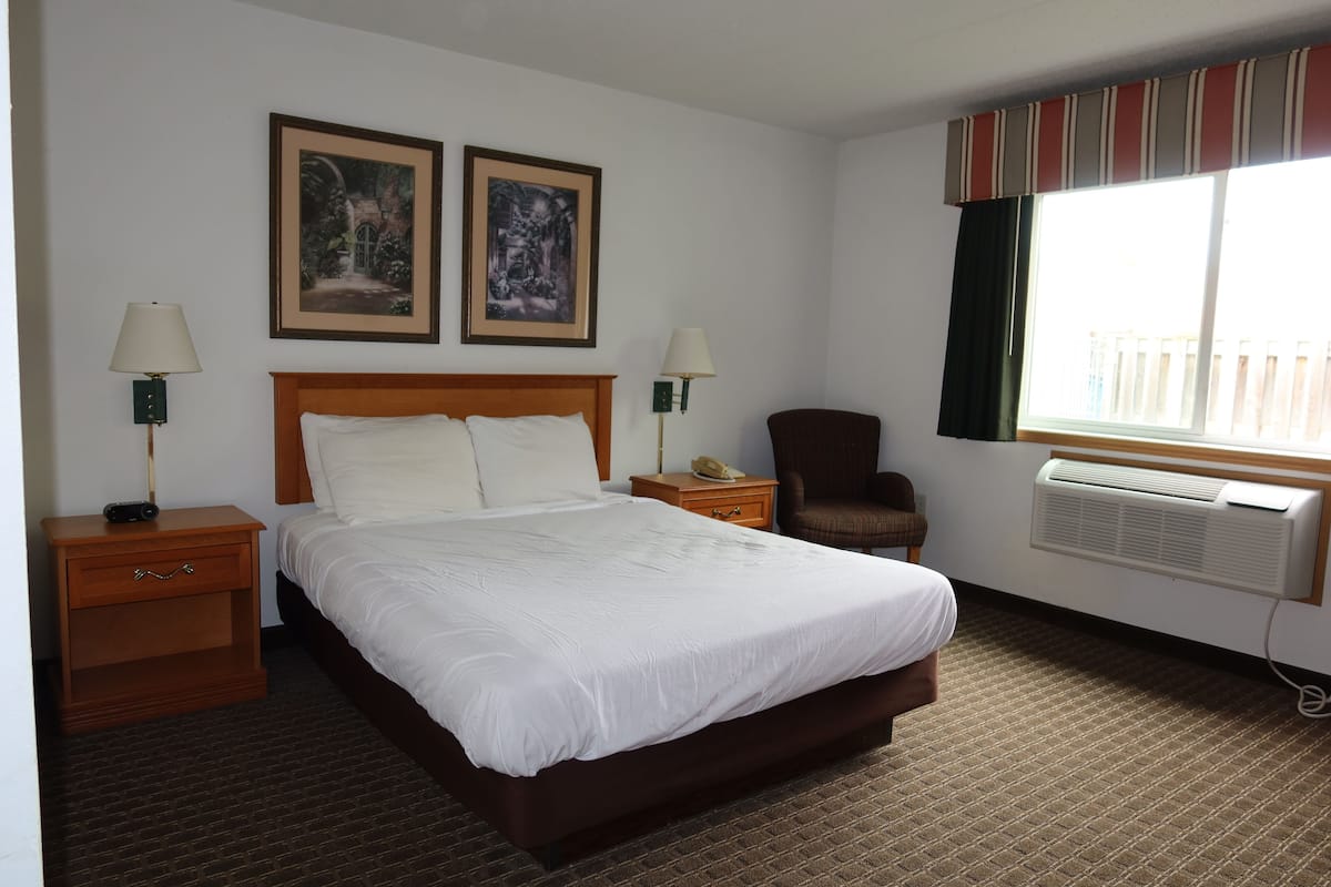 Una habitación de hotel con una cama grande, dos mesitas de noche con lámparas, una silla, una ventana con cortinas y dos cuadros enmarcados en la pared.