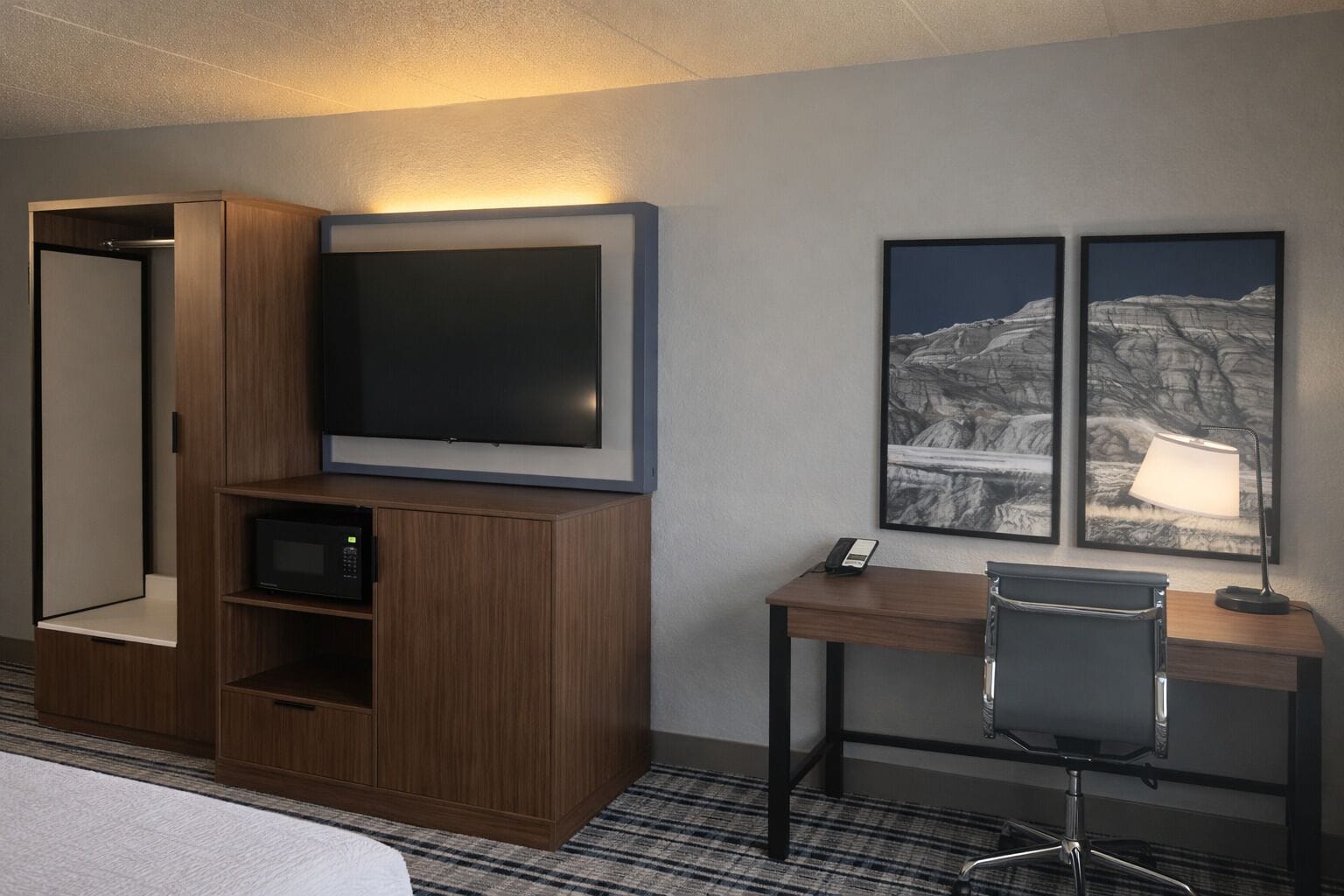 A hotel room with a bed, a TV, a desk with a chair, and two framed pictures on the wall.