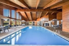 A modern indoor swimming pool with wooden beams, clear windows, and sun loungers.