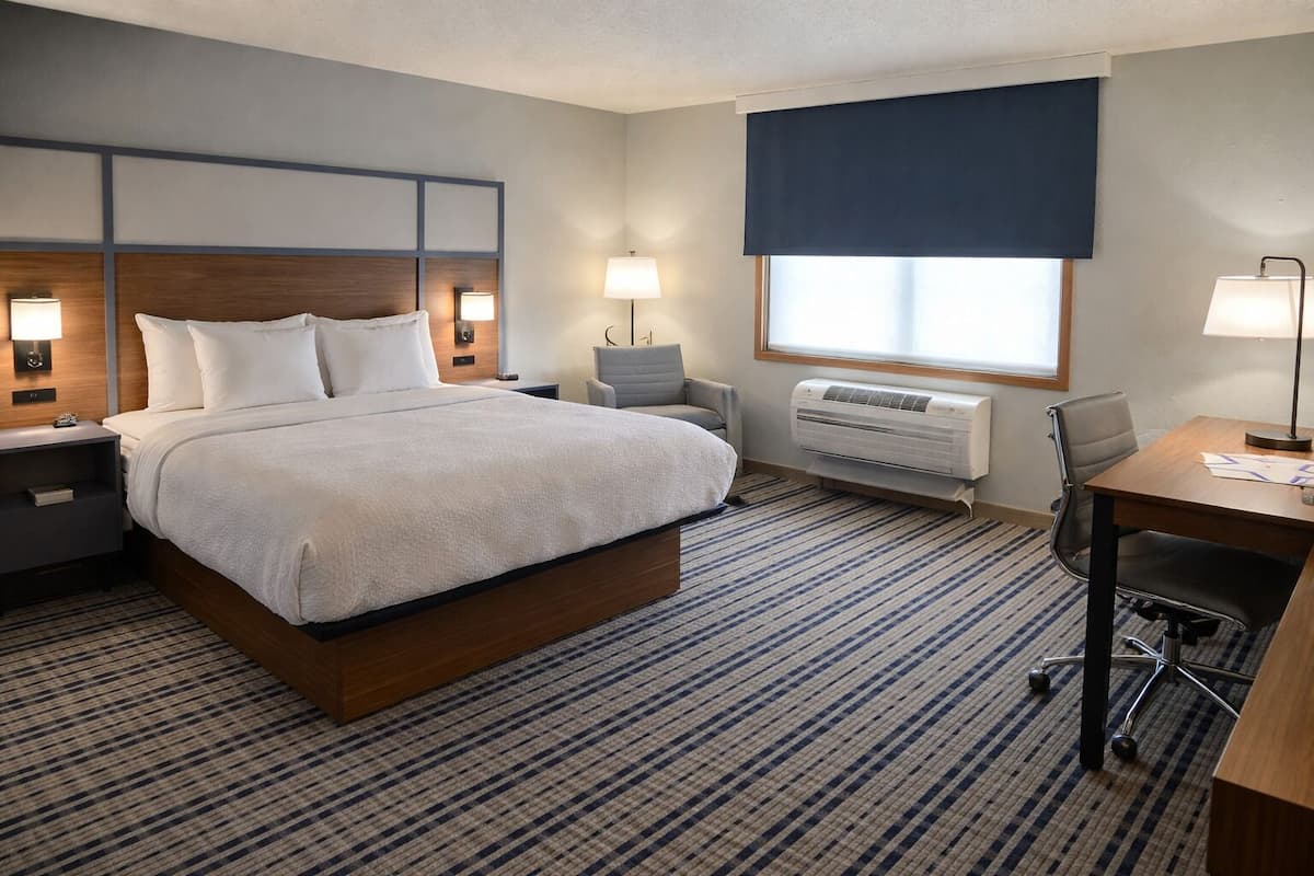 A hotel room with a large bed, bedside tables with lamps, a desk with a chair, a window with a dark blind, and an air conditioning unit.
