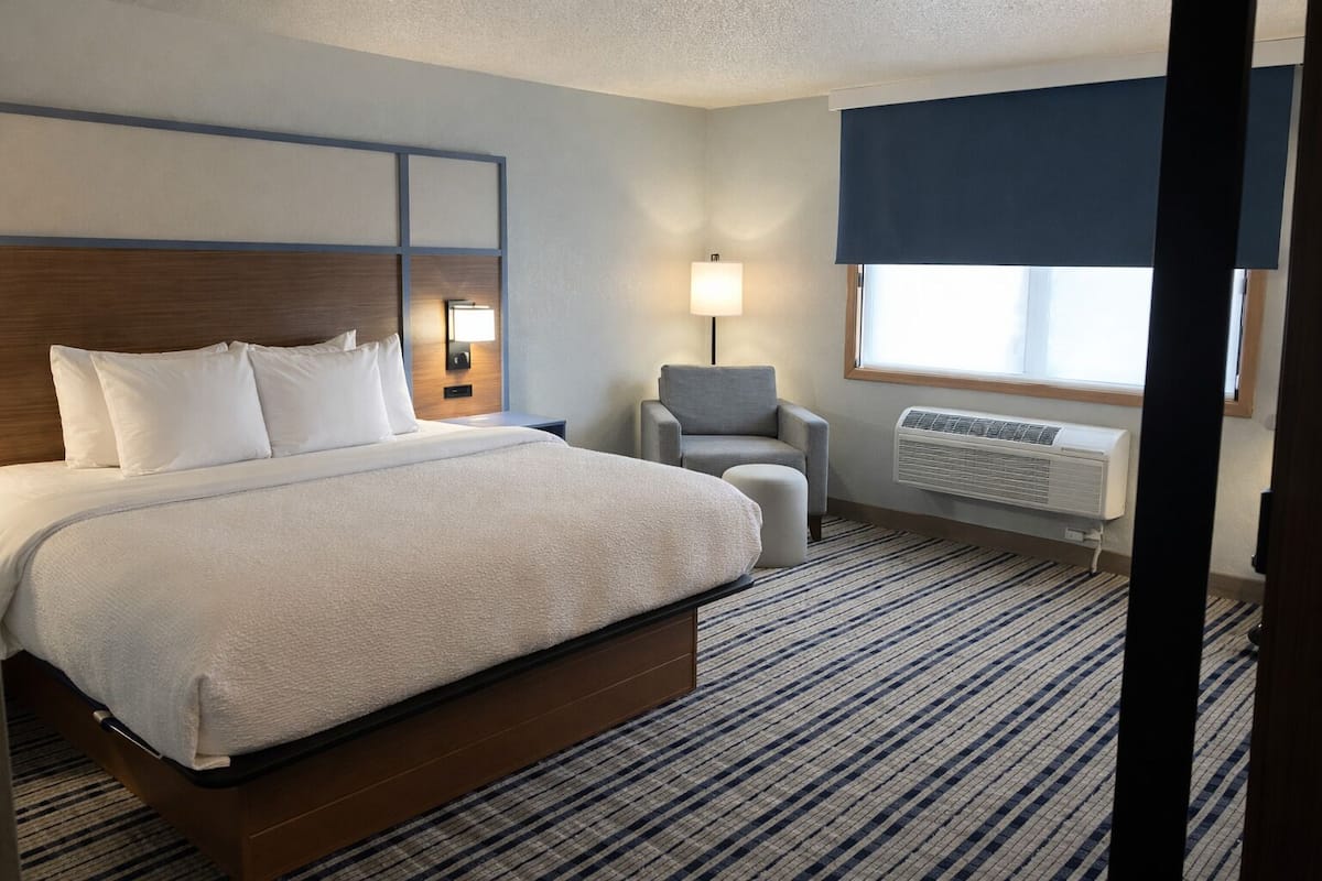A hotel room with a large bed, a chair, a desk with a lamp, and a window with blinds.