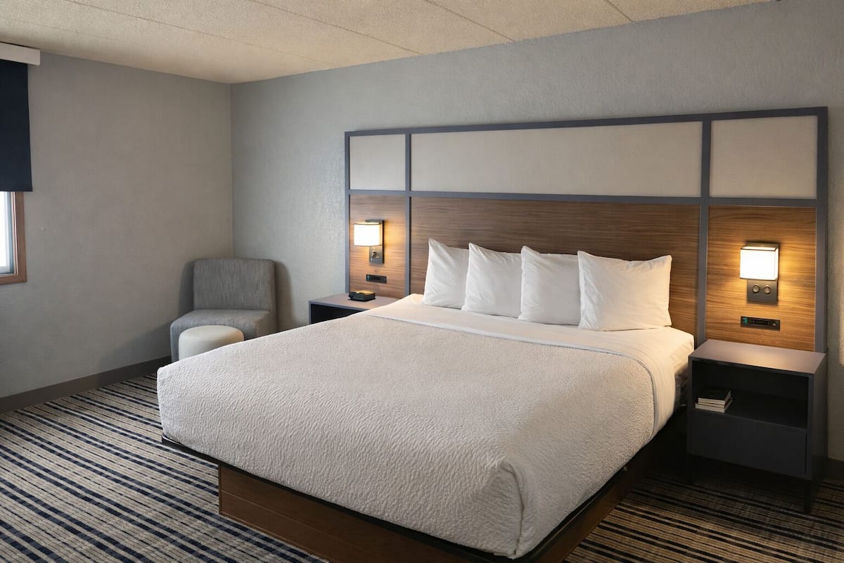 A hotel room with a large bed, a chair, a nightstand with a lamp, and a wall-mounted control panel.