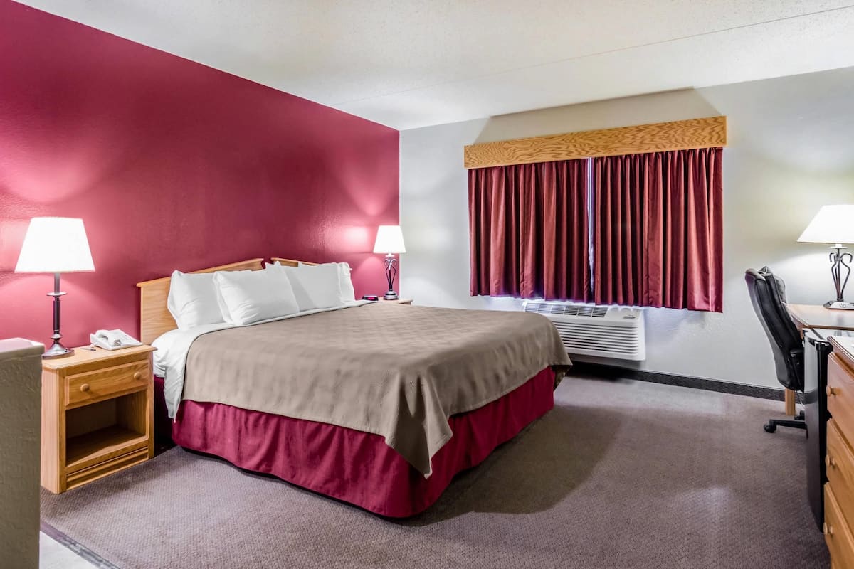 Une chambre d’hôtel avec un lit, des tables de chevet, des lampes, un bureau et une fenêtre avec des rideaux.
