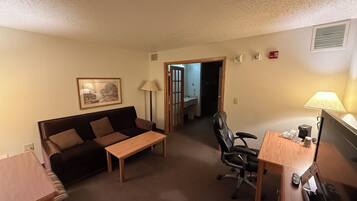 Un espace de vie compact comprenant un canapé, une petite table, un bureau avec une chaise, une lampe et un téléviseur. Une porte donne accès à la salle de bain.