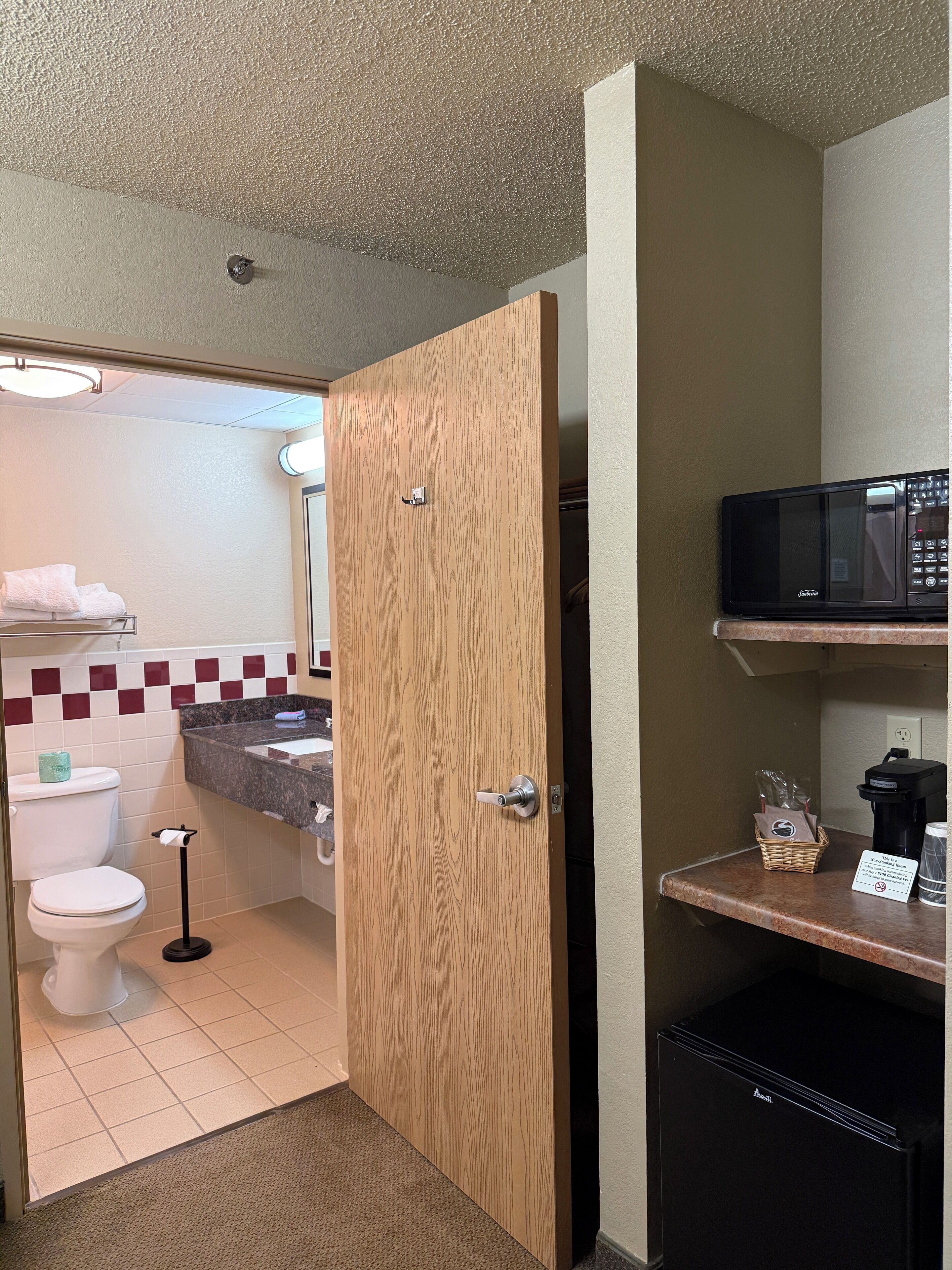 Een kleine hotelbadkamer met een houten deur, een wit toilet, een wastafel met handdoekhouder en een magnetron op een plank.
