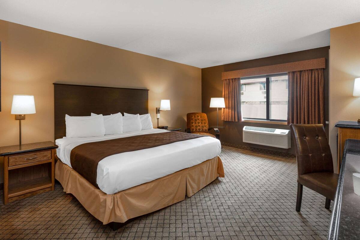 A hotel room with a large bed, two bedside tables, a chair, a window with curtains, and an air conditioning unit.