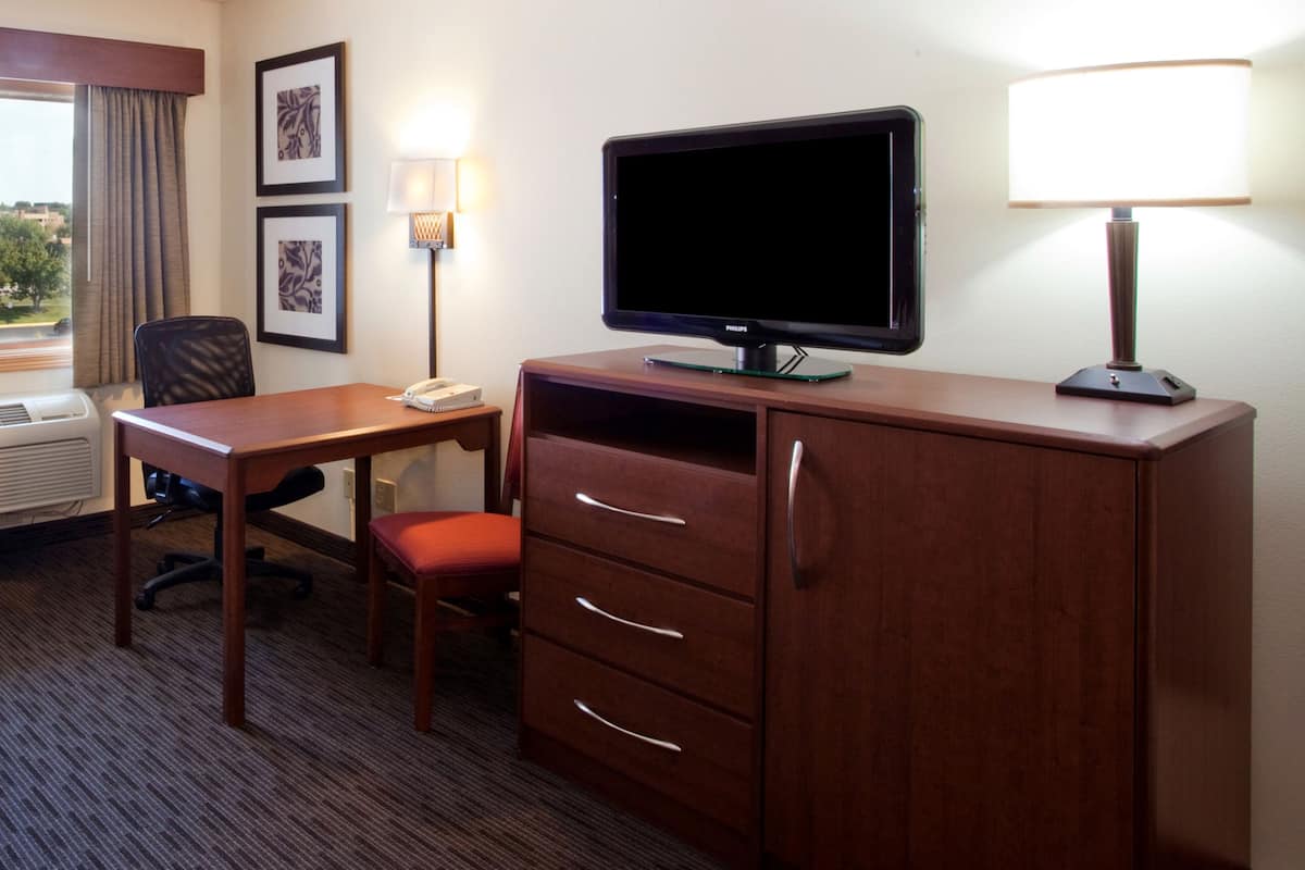 A hotel room with a wooden desk, a television, a lamp, a chair, and a dresser with drawers.