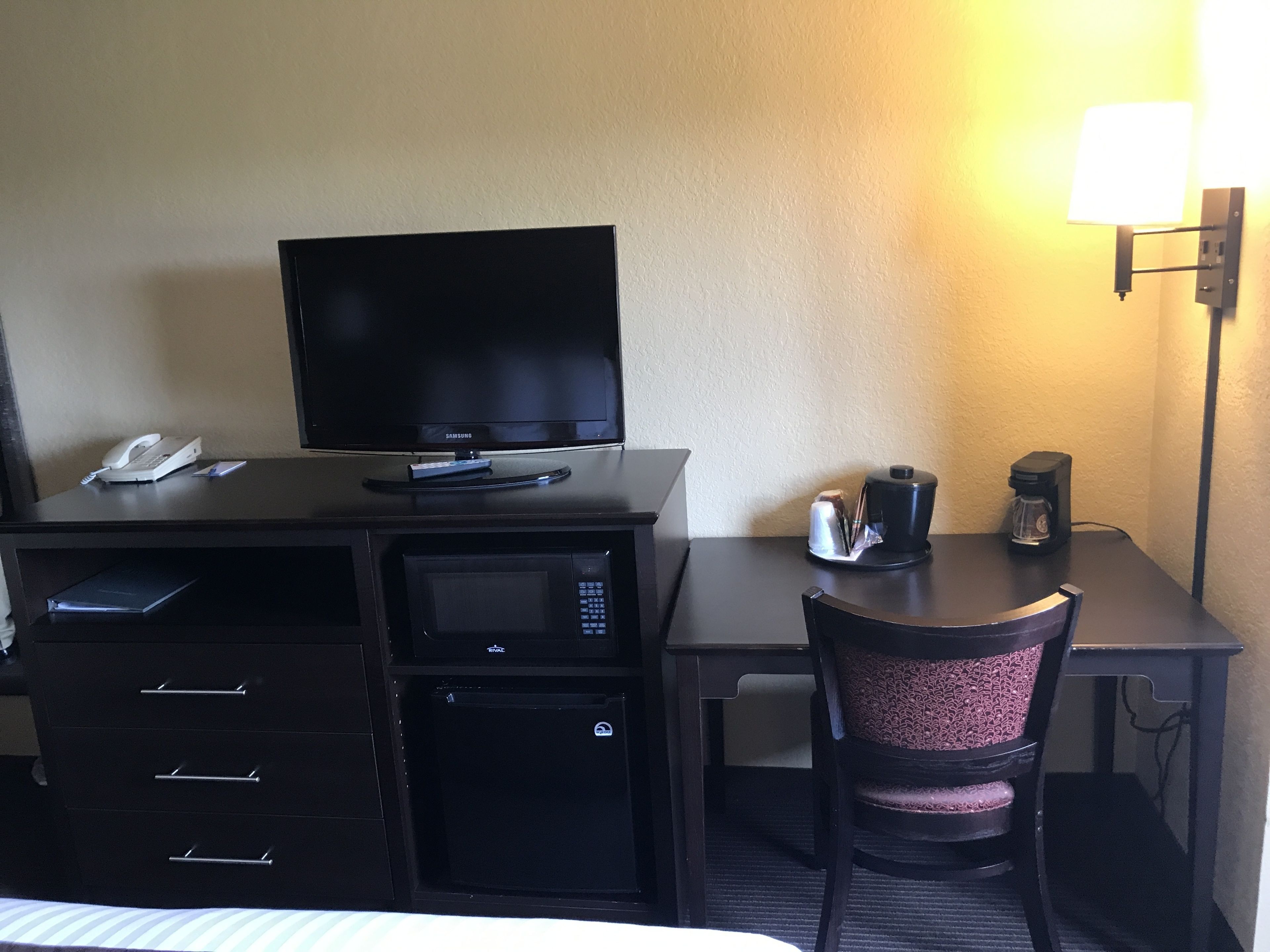 Quarto de hotel com escrivaninha, televisão, micro-ondas, máquina de café e uma cadeira.
