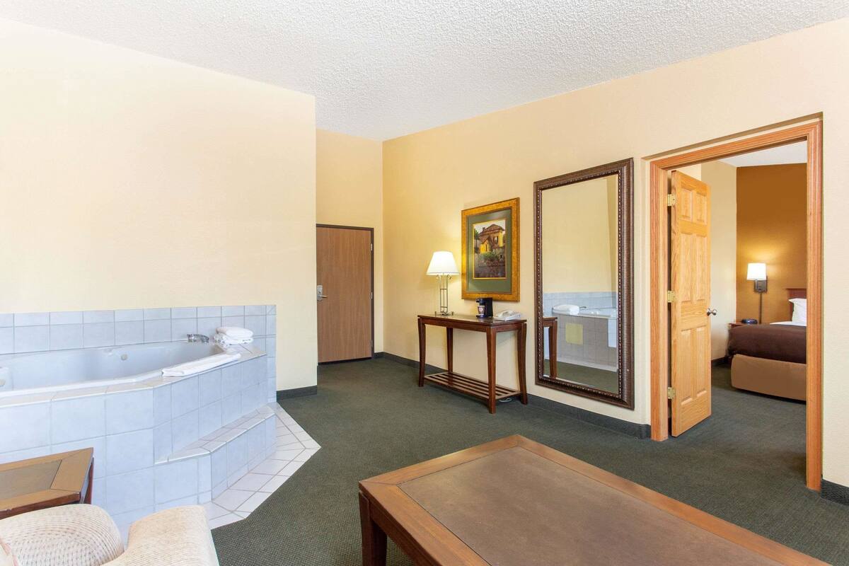 A hotel bathroom with a bathtub, a mirror, a table, and a door leading to a bedroom.