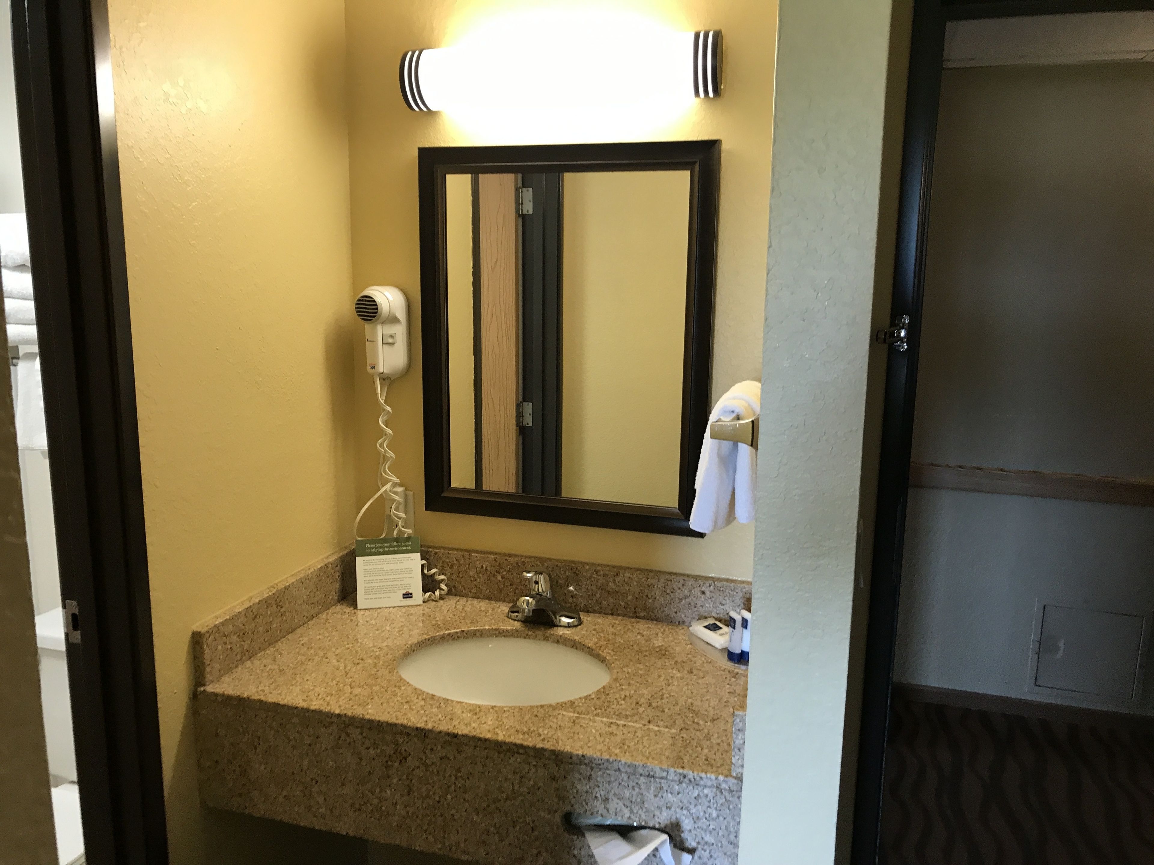 Baño de hotel con lavamanos de encimera de granito, espejo, secador de pelo y toalla colgada en la pared.
