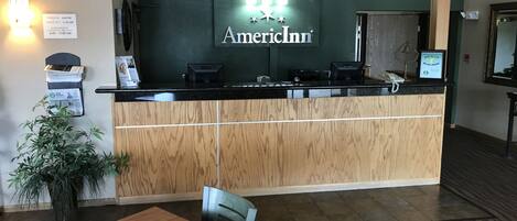 Una reception con una insegna che riporta "AmericInn" e un orario di check-in/out. A sinistra, una pianta e, in primo piano, un tavolo di legno con una sedia.