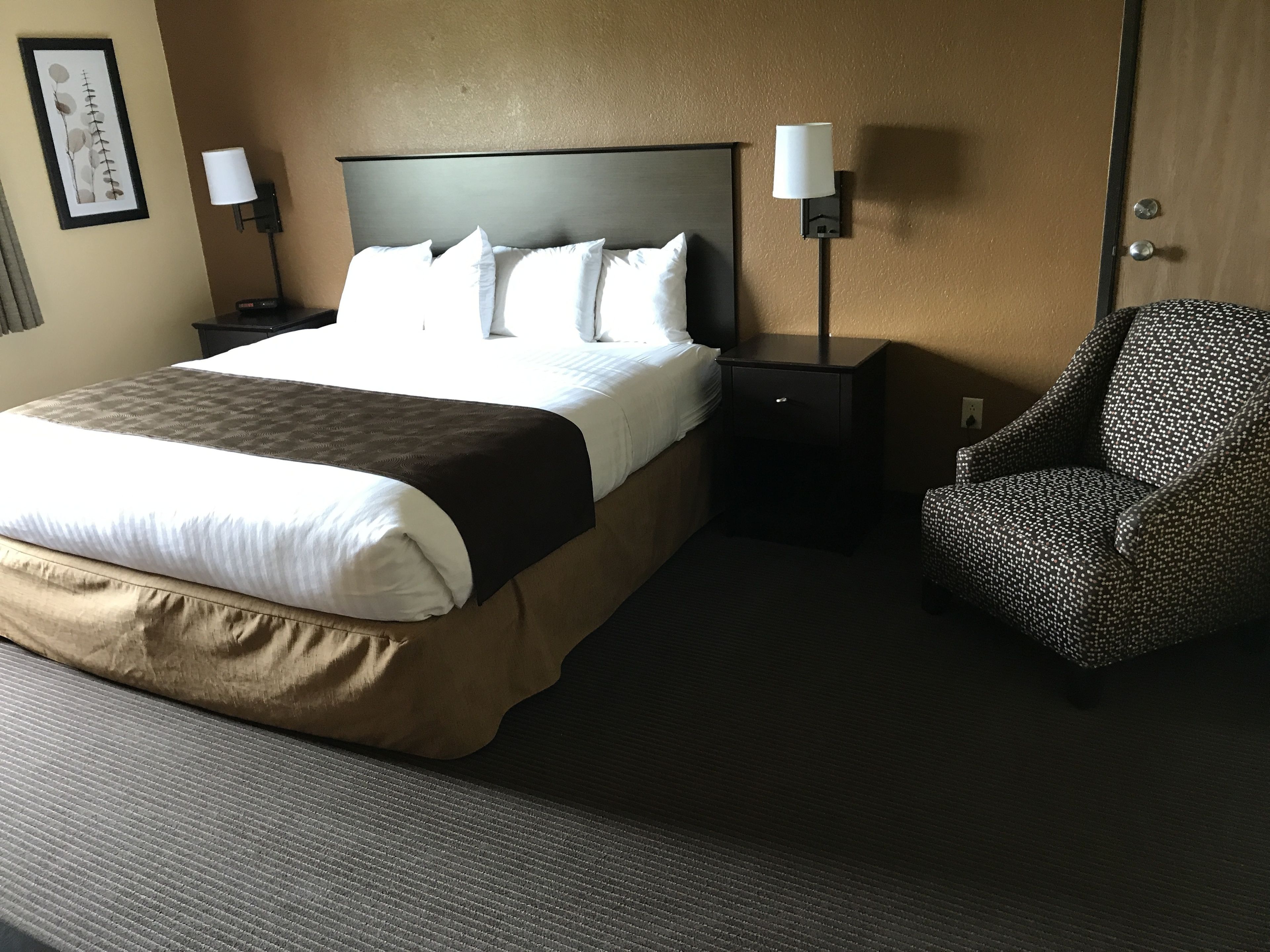 A neatly made bed with a headboard, two bedside tables with lamps, a patterned armchair, and a framed picture on the wall.