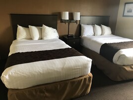 A hotel room with two beds, each with white bedding and brown throw pillows. There are two bedside tables with lamps, and a framed picture on the wall.
