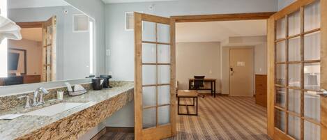Une chambre d’hôtel dotée d’un lavabo avec un plan en granit, d’un grand miroir, d’une porte coulissante à cadre en bois, d’une moquette à rayures et d’une petite table avec des chaises.