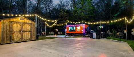 Une structure en bois avec une porte de garage, un camion-restaurant illuminé de néons et une zone pavée décorée de guirlandes lumineuses.