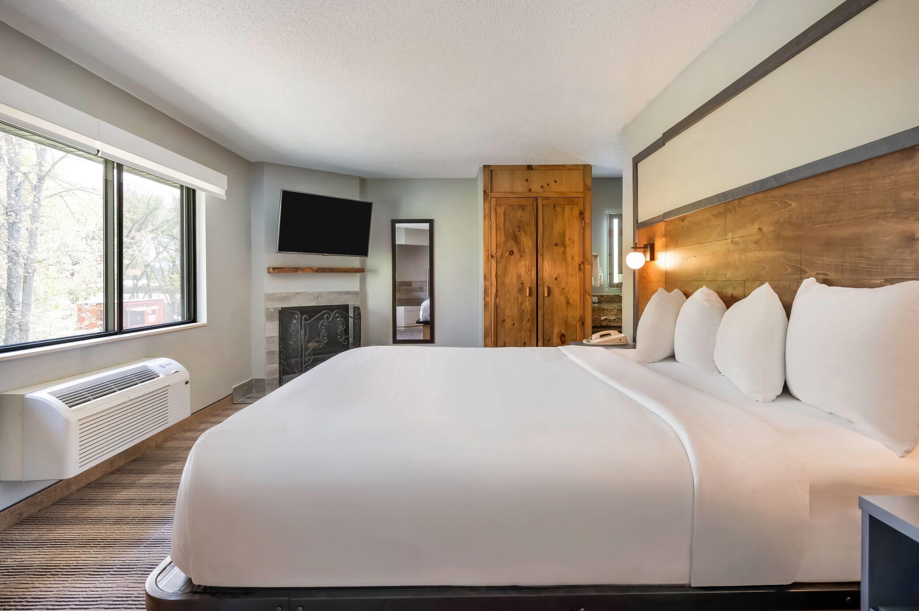 A large bed with white bedding, a mounted TV, a wooden wardrobe, and a fireplace.