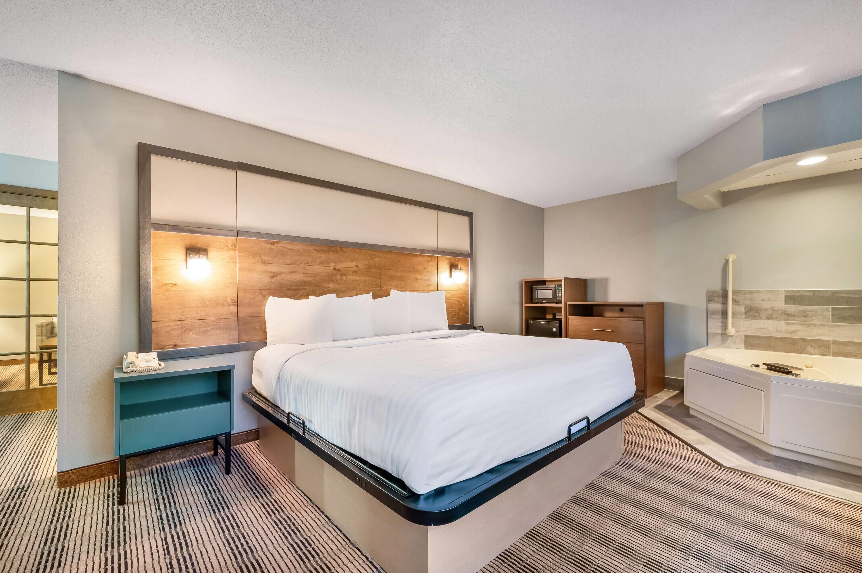 A hotel room with a bed, bedside table, nightstand, and a bathroom with a bathtub.