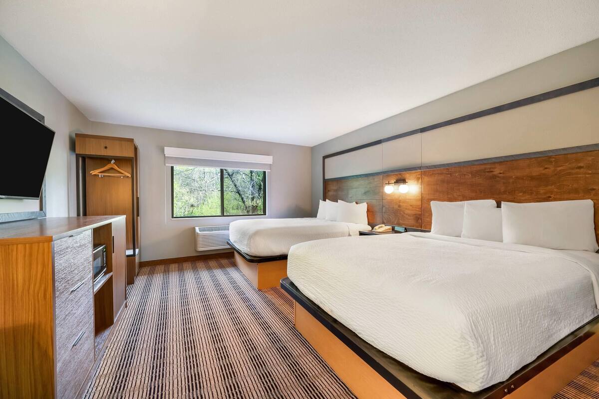 A hotel room with a bed, a desk, a TV, a window, and a wooden headboard.