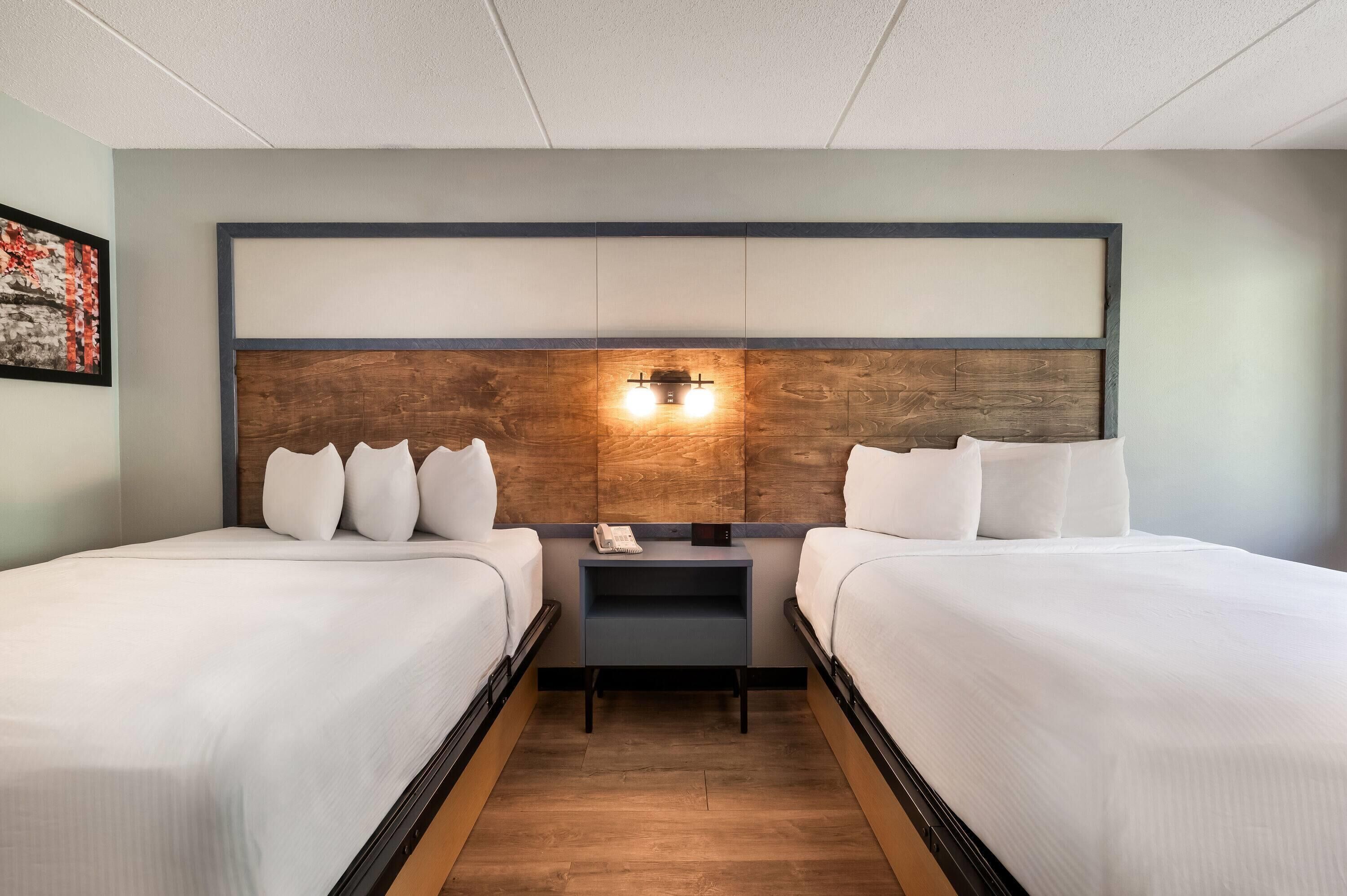 A hotel room with two beds, a wooden headboard, a nightstand with a phone, and a wall-mounted light.