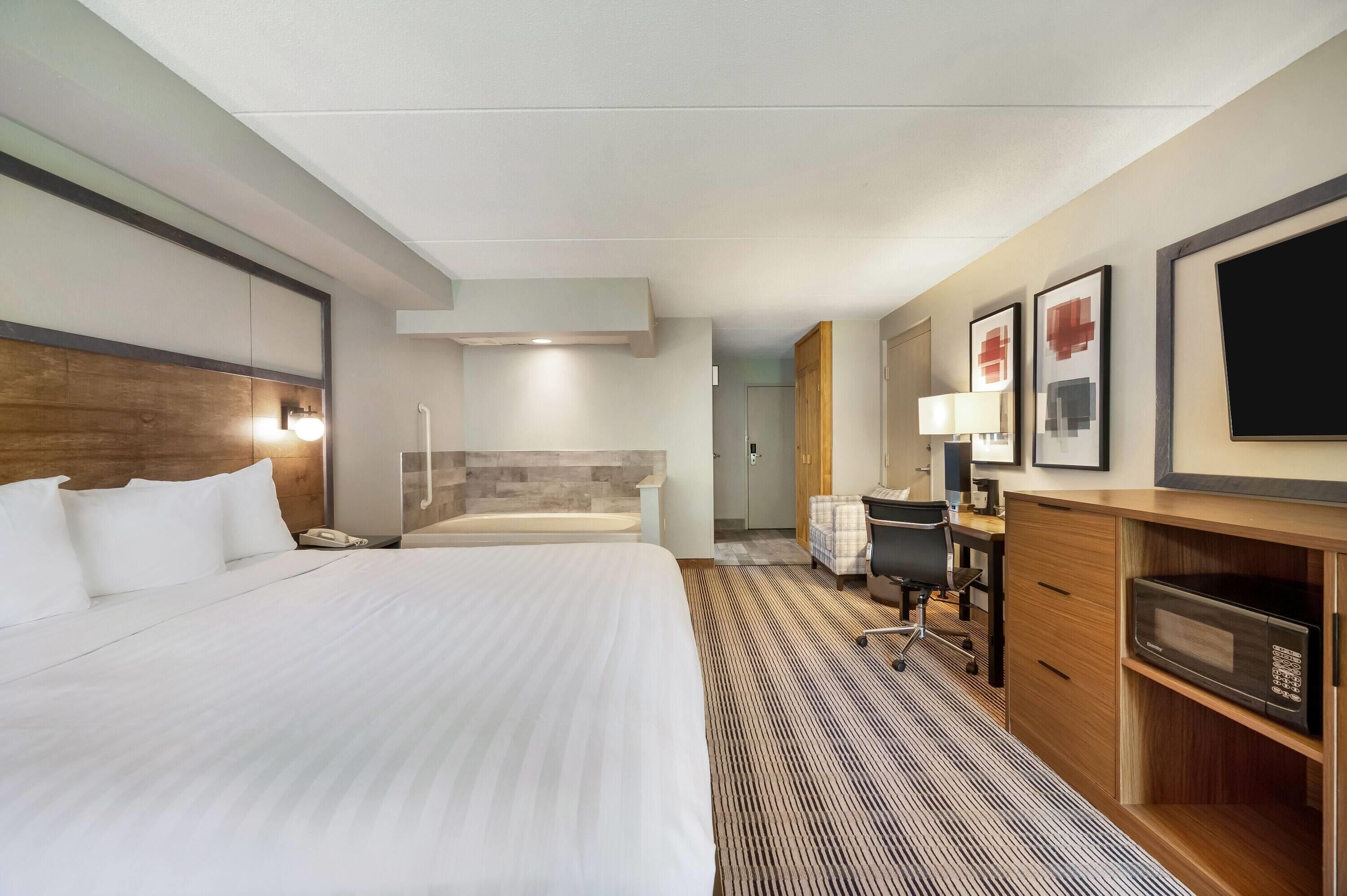 A hotel room with a large bed, a desk with a chair, a microwave, a TV, and a bathroom with a bathtub.