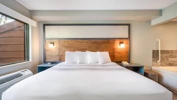 Habitación de hotel con una cama grande, mesitas de noche, una mesita auxiliar, un televisor montado en la pared, una ventana y un baño con bañera.