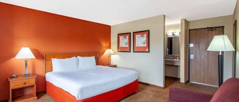 Een hotelkamer met een bed, twee bedlampen, een rode stoel, een badkamer met een spiegel en twee ingelijste foto's aan de muur.